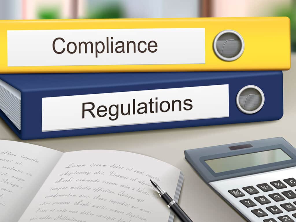 Yellow and blue binders labeled "Compliance" and "Regulations" on a desk with a pen, open book, and calculator, in an office setting.