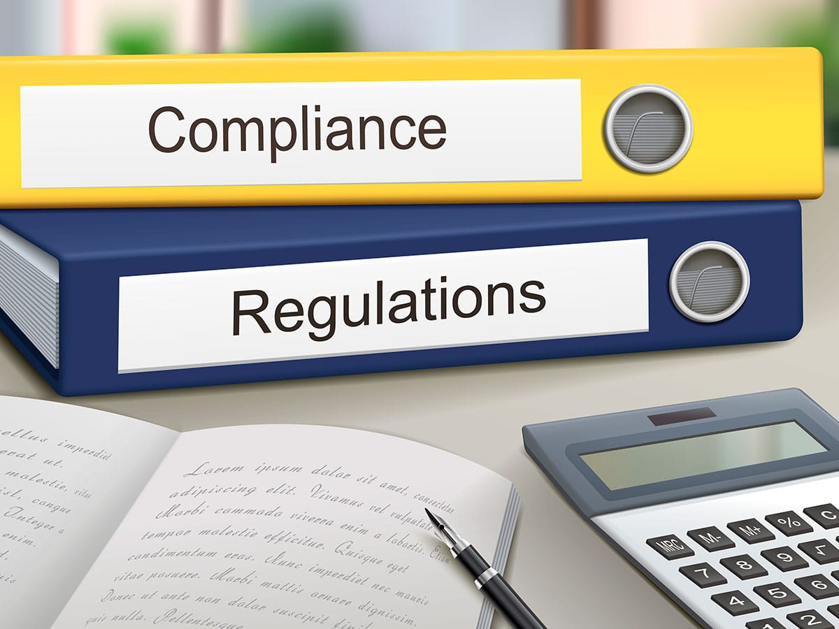 Yellow and blue binders labeled "Compliance" and "Regulations" on a desk with a pen, open book, and calculator, in an office setting.