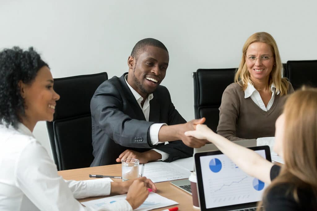Business meeting with four people around a table. A man in a suit and a woman shake hands, smiling. A laptop shows graphs.