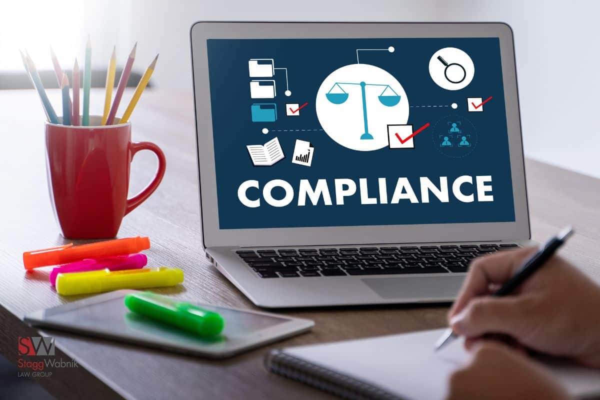 Laptop showing "COMPLIANCE" with icons; person writing notes. Red cup with pencils and colorful markers on desk. Office setting.