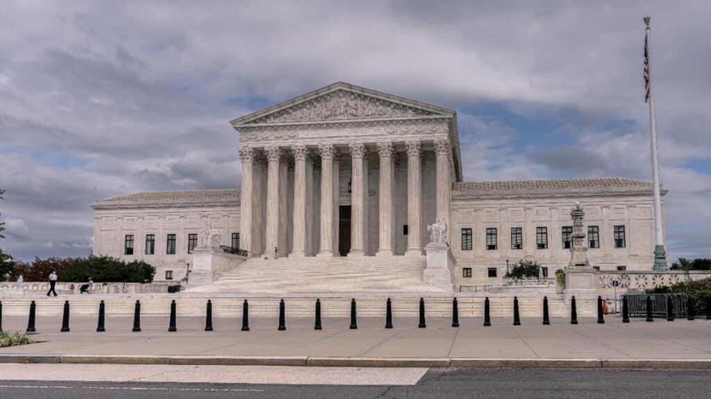 U.S. Supreme Court building with columns and steps, a few people walking, cloudy sky, and American flag on a pole.