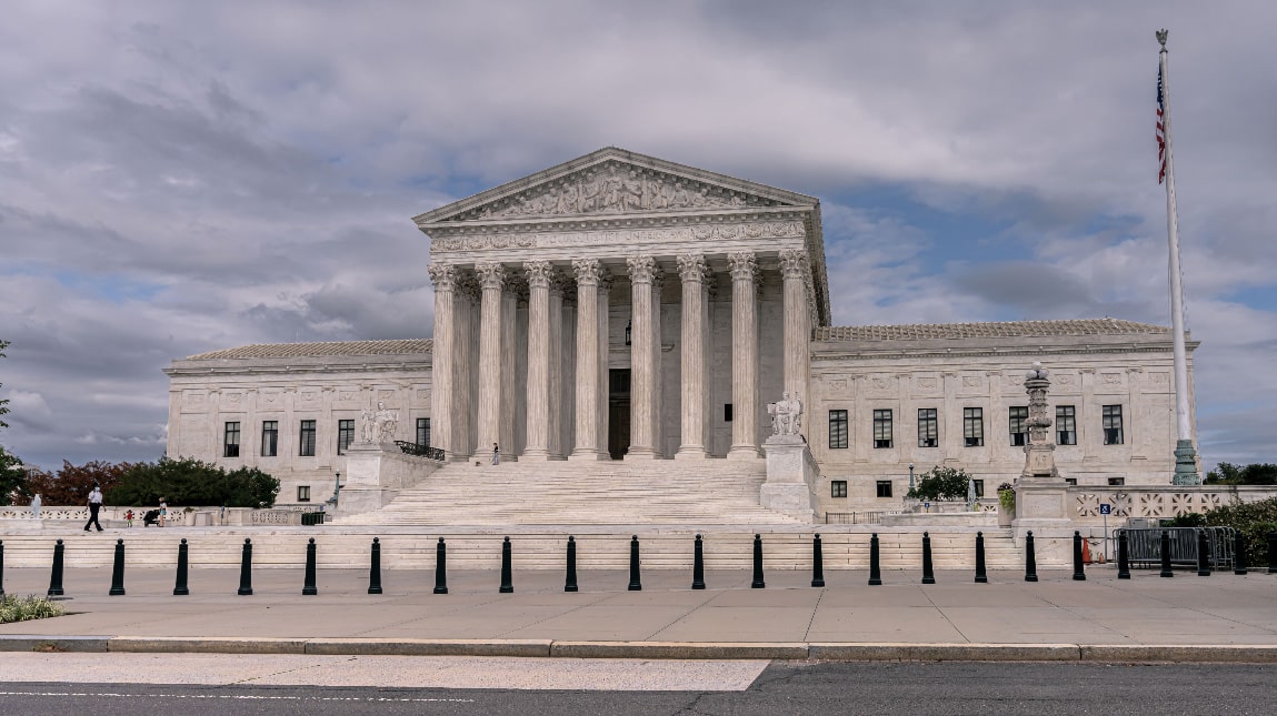 U.S. Supreme Court building with columns and steps, a few people walking, cloudy sky, and American flag on a pole.