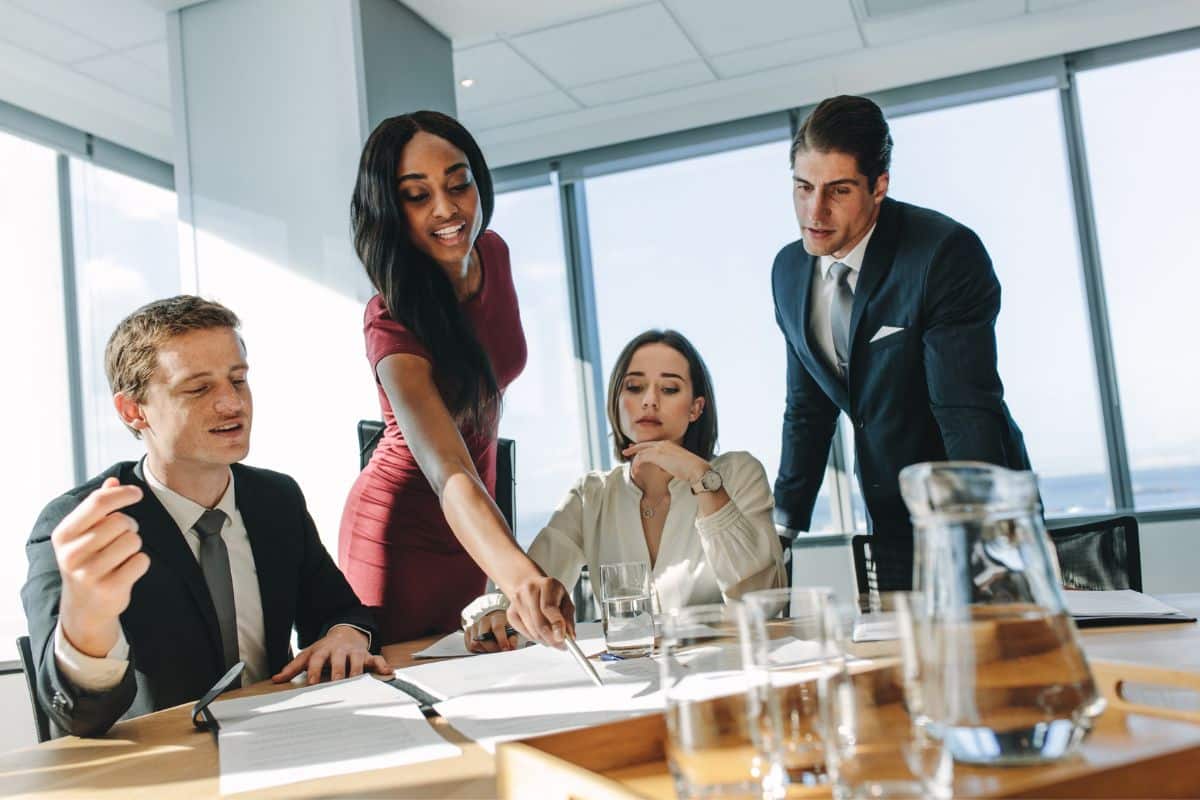 Four people in a bright office, reviewing documents. One woman points at papers, others listen attentively. Glasses and jar on the table. Business attire.